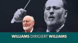 Komponist und Dirigent John Williams vor einem Plakat, welches ihn dirigierend zeigt